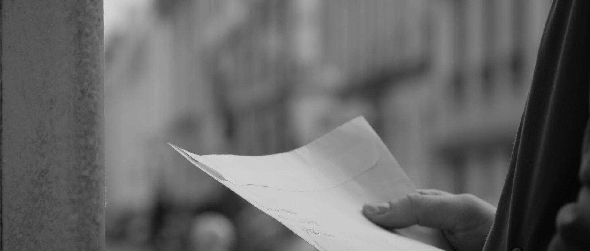 Main qui tient une lettre pliée, façades parisiennes floues à l'arrière-plan. Plan rapproché, noir et blanc.