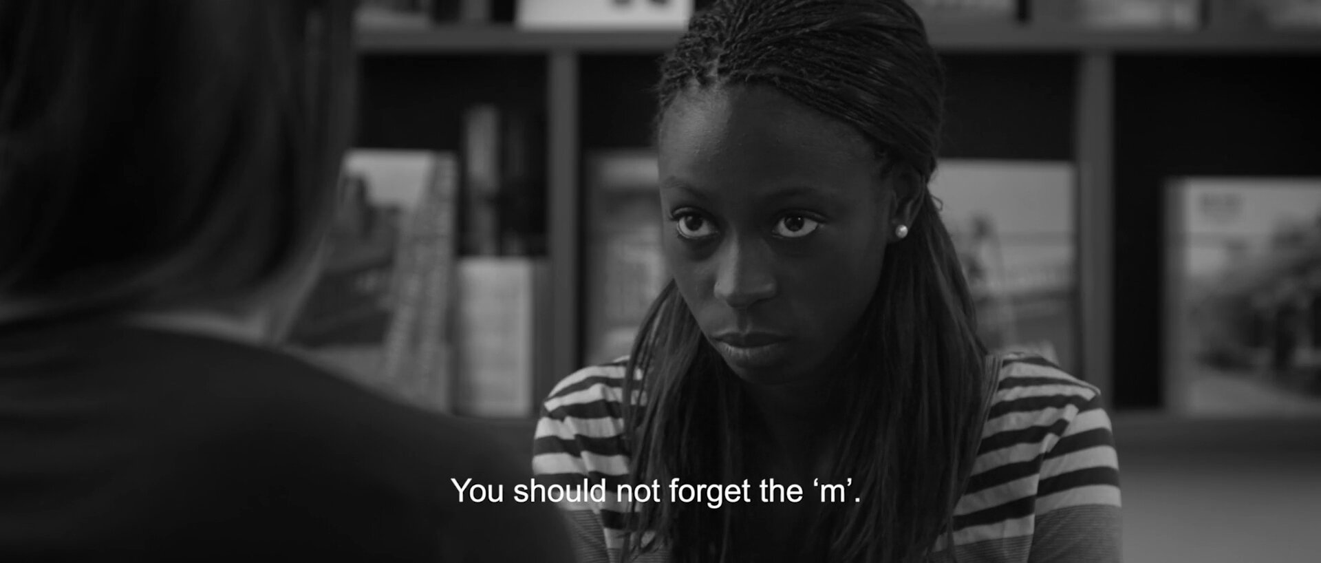 Portrait face caméra d'une jeune femme dans une bibliothèque, sous-titre 'You should not forget the m.' en bas du cadre. Noir et blanc, scope.